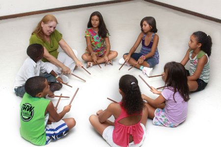 Porfa. Carmen Mettig setada ao chão numa roda de crianças, realizando uma atividade rítmica com baquetas de bateria